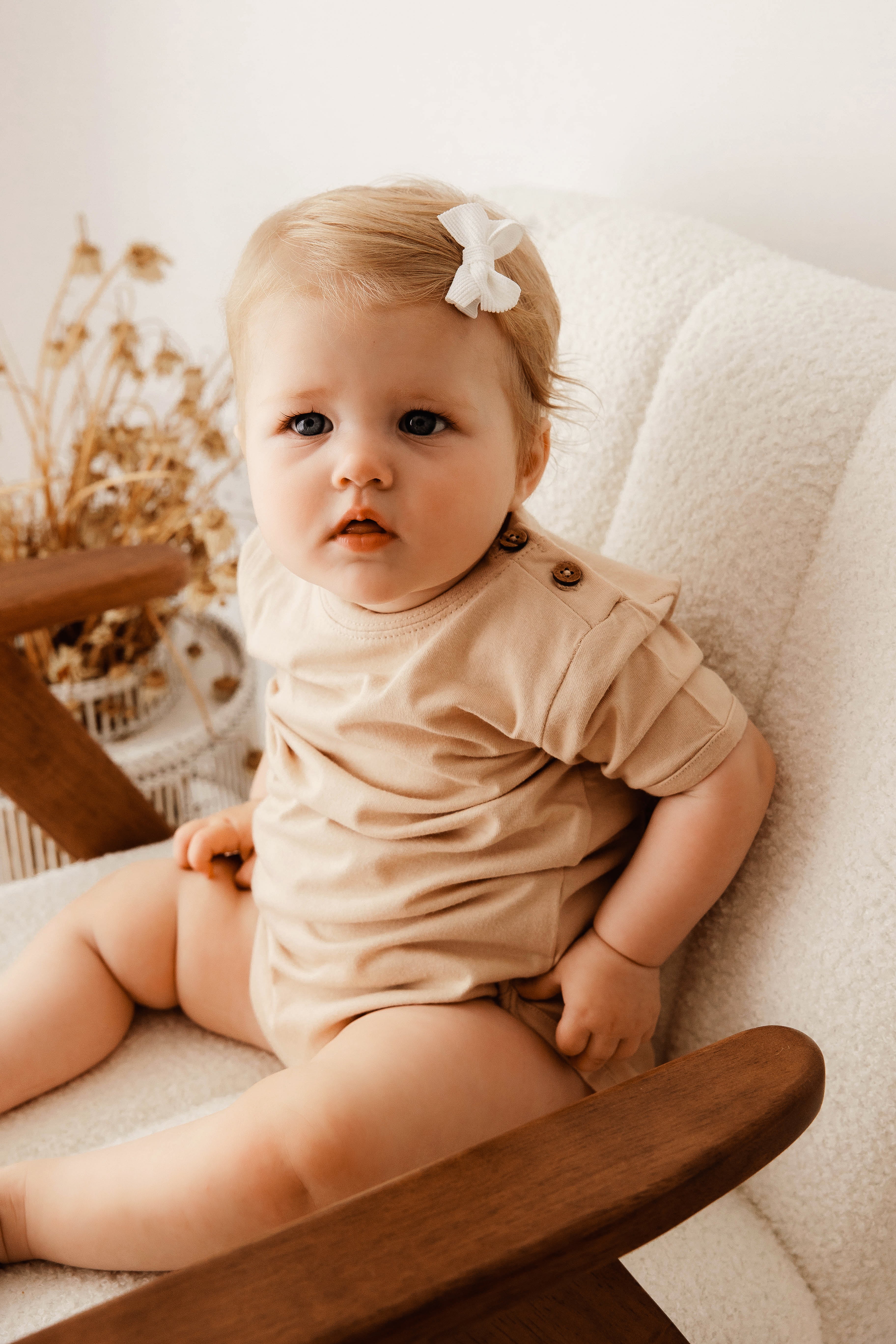 Baby sitting on a wooden chair wearing a beige outfit with a white bow in hair.