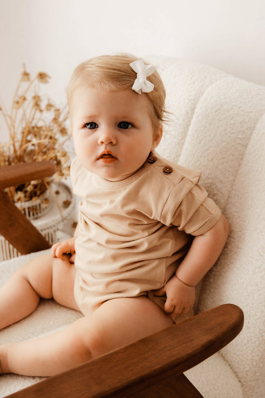 Baby sitting on a wooden chair wearing a beige outfit with a white bow in hair.